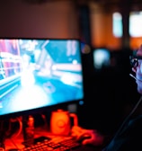 A person wearing headphones is sitting in front of a computer monitor. The screen displays a vibrant, illuminated video game scene, while the person appears focused on gaming. The ambient lighting is dark with red and blue tones, and a backlight from the monitor highlights the headphones.