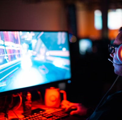 A person wearing headphones is sitting in front of a computer monitor. The screen displays a vibrant, illuminated video game scene, while the person appears focused on gaming. The ambient lighting is dark with red and blue tones, and a backlight from the monitor highlights the headphones.