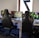 A row of gaming stations equipped with large monitors, gaming chairs, keyboards, and headphones. The setup is organized and ready for gaming, featuring a colorful screen display. The room has a modern and tech-savvy ambiance.
