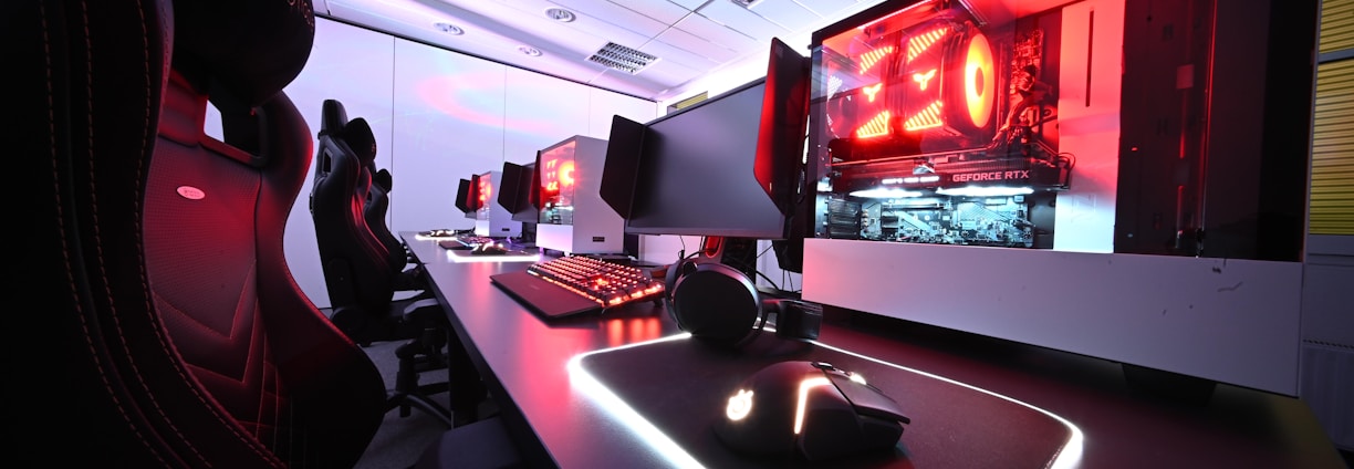 A row of high-end gaming computers on sleek desks, featuring RGB lighting and gaming accessories. The computers are housed in transparent cases with visible components, emitting a red glow. Comfortable gaming chairs with ergonomic design are positioned next to the desks, and the room's ceiling is modern with recessed lighting.
