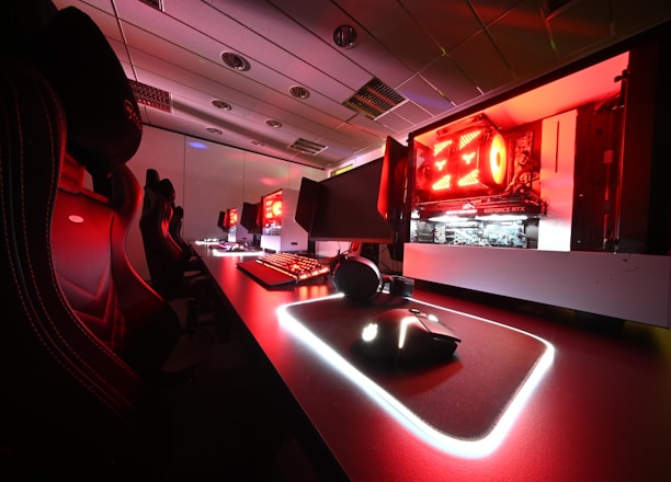 A modern gaming setup featuring several high-end computers with illuminated red interior lighting. The room is equipped with gaming chairs exhibiting a sleek black and red design. Multiple monitors are arranged on a long desk, and peripheral devices like keyboards and mice are uniformly placed, reflecting an ambiance ideal for gaming or competitive play.