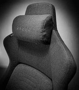 A high-quality, fabric gaming chair featuring ergonomic design and enhanced comfort. The chair has a distinct headrest with visible stitching, embossed branding, and a modern aesthetic.