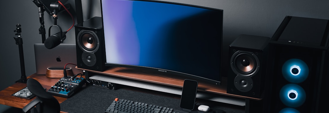 A modern gaming and streaming setup with a curved monitor placed centrally on a wooden desk. Surrounding the monitor are powerful speakers and a high-end PC tower with glowing fans. Various peripherals, including a mechanical keyboard, ergonomic mouse, and audio mixer, are neatly arranged. Overhead, a professional microphone and stand are ready for use, possibly for gaming commentary or podcasting.
